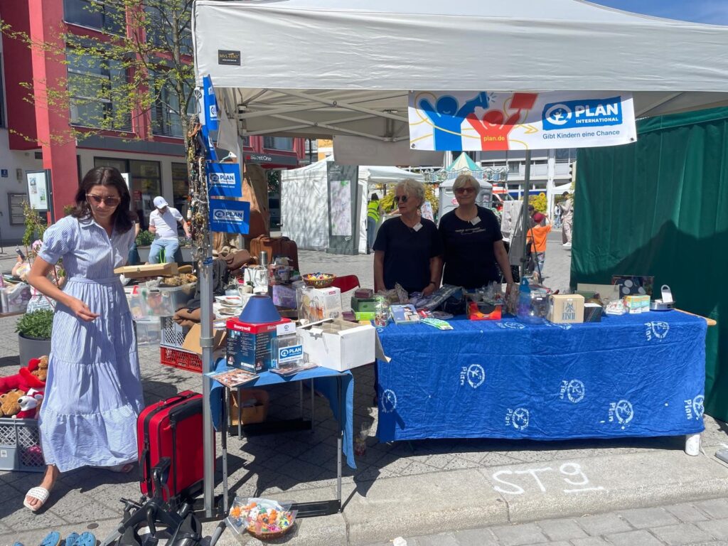 Three women stand at an outdoor market stall under a white tent, displaying various items for sale. The table and banners feature the PLAN International logo. One woman arranges items, while the others stand behind the table.