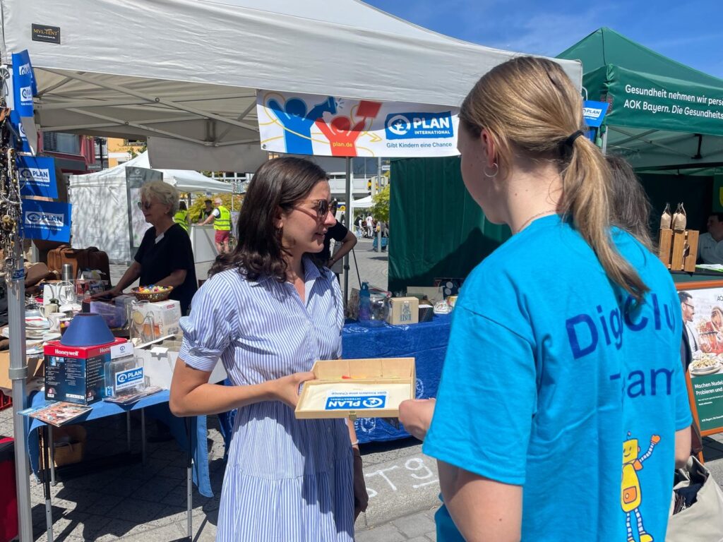 Two young women talk in front of a booth at an outdoor fair. One holds a tray and wears sunglasses; the other wears a blue shirt with Plan on the back. A white tent and informational banners are visible behind them.