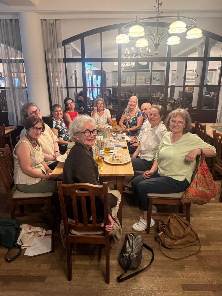 Eine Gruppe von elf Erwachsenen sitzt in einem Restaurant um einen Holztisch herum und lächelt in die Kamera. Auf dem Tisch stehen Teller, Gläser und Getränke. Die Atmosphäre ist ungezwungen und die Beleuchtung warm.