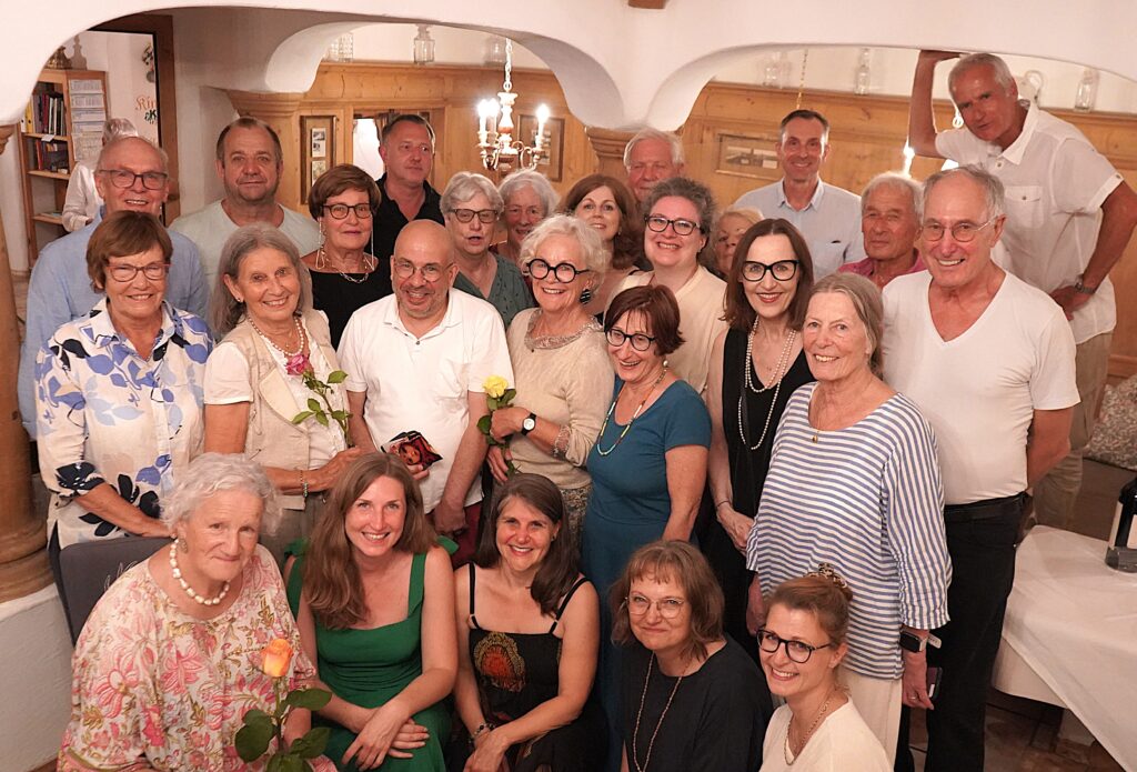 Eine Gruppe lächelnder Erwachsener und Senioren, von denen einige Rosen in den Händen halten, posiert gemeinsam in einem warm beleuchteten Raum mit Holzdekor.