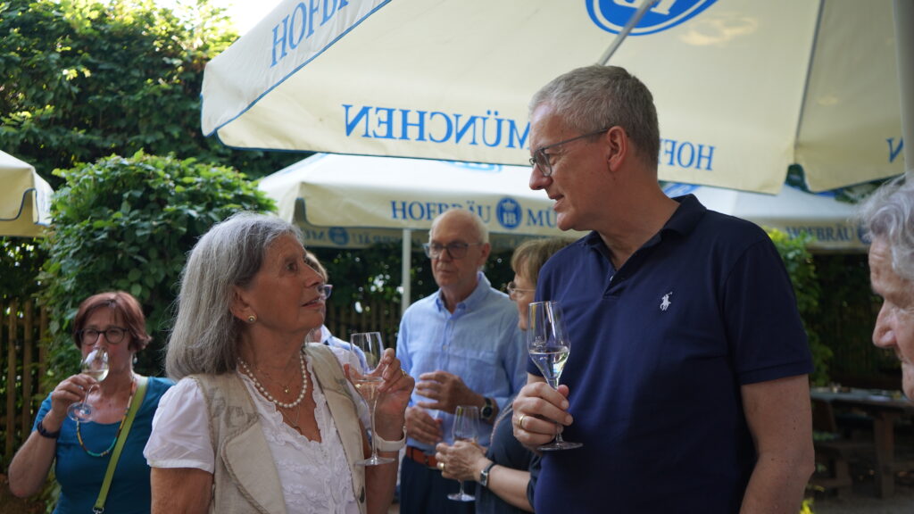 Eine Gruppe von Menschen steht im Freien, hält Getränke in der Hand und unterhält sich unter großen weißen Sonnenschirmen mit dem Aufdruck „Hofbräu München“. Zwei Personen im Vordergrund unterhalten sich und lächeln. Es ist ein entspanntes Beisammensein.
