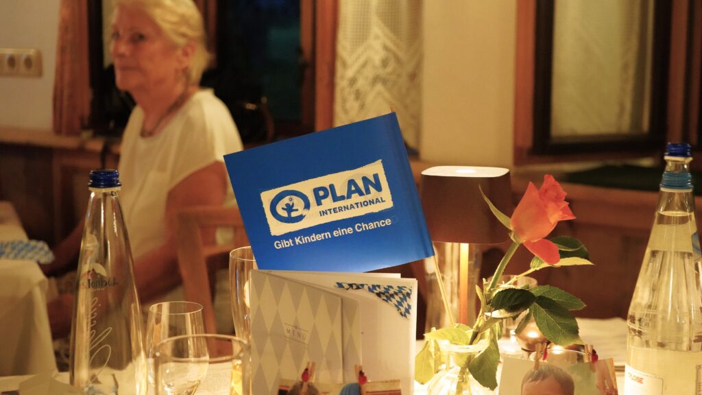 Ein Tisch mit einer Rose, Wasserflaschen, Gläsern und einer blauen Broschüre von Plan International mit der Aufschrift „Gibt Kindern eine Chance“. Im Hintergrund sitzt eine Frau in einem warm beleuchteten Raum.
