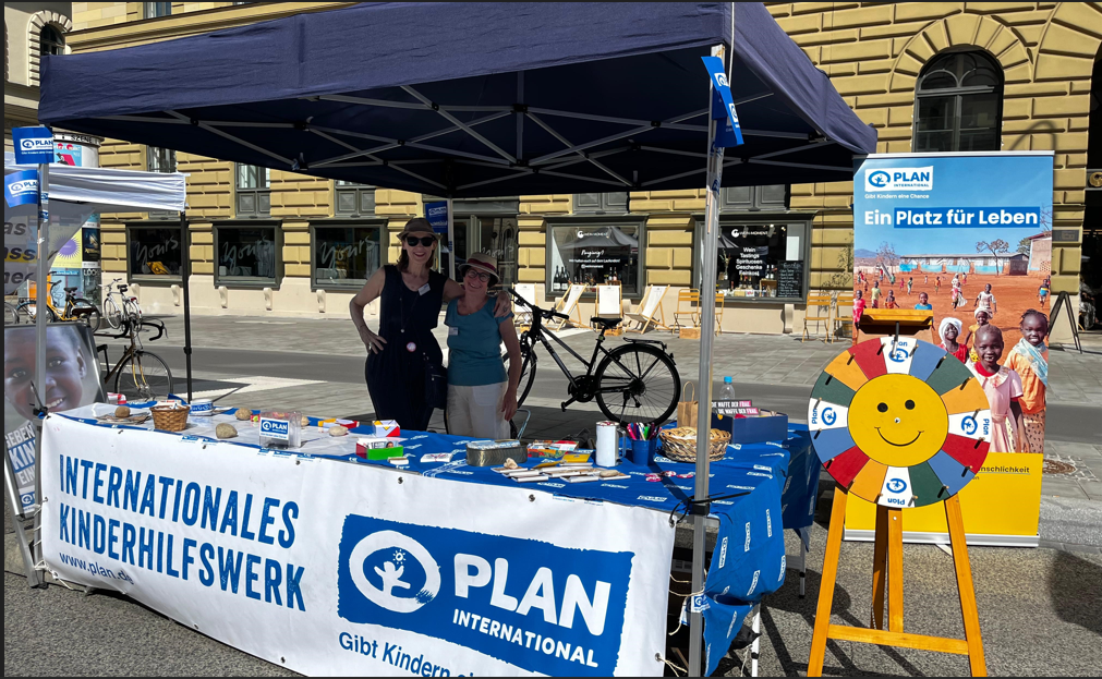 Zwei Personen stehen unter einem blauen Zelt am Stand von Plan International auf einer sonnigen Straße, umgeben von Flyern, einem Glücksrad und Bannern, die für Kinderhilfe werben. Im Hintergrund sind ein Fahrrad und ein historisches Gebäude zu sehen.