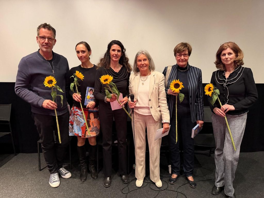 Sechs Personen stehen in einer Reihe auf einer Bühne und halten jeweils eine Sonnenblume in der Hand.