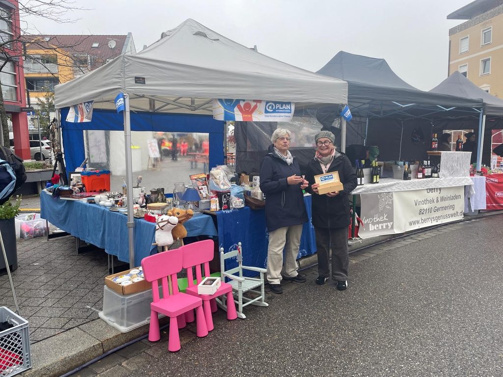 Zwei Frauen stehen vor einem Flohmarktstand, auf dem verschiedene Haushaltsgegenstände, Spielzeug und ein Schild von Plan International ausgestellt sind.