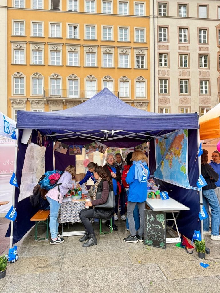 Bei einer Veranstaltung im Freien vor einem hohen gelben Gebäude versammeln sich Menschen unter einem blauen Pop-up-Vordach, an dem ein Schild mit der Aufschrift Wir passen, Bäume wachsen steht.