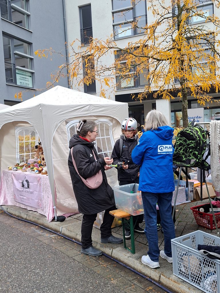 Zwei Frauen und ein Kind mit Helm stehen an einem Marktstand im Freien, auf dem Stofftiere ausgestellt sind.