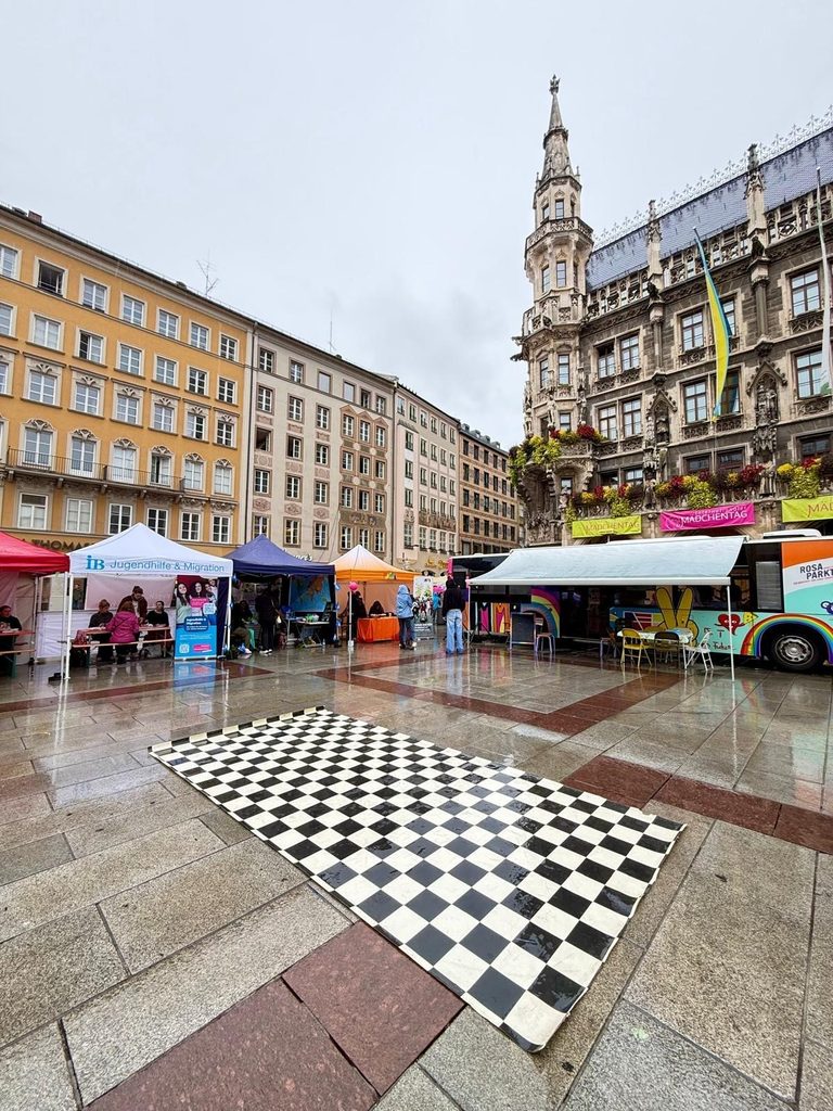 Auf dem nassen Pflaster eines Stadtplatzes, umgeben von bunten Verkaufszelten und einem geschmückten Bus in der Nähe eines historischen Gebäudes, liegt eine karierte Fußmatte.